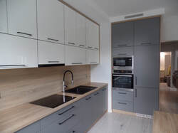 Nowoczesna kuchnia z szarymi dolnymi szafkami i białymi górnymi, drewniany blat, czarny zlew i płyta indukcyjna, wysoka zabudowa z piekarnikiem i mikrofalówką.