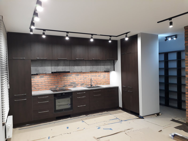 Nowoczesna kuchnia z ciemnymi, drewnopodobnymi frontami szafek, ceglaną ścianą nad blatem i oświetleniem szynowym, widoczne kartony na podłodze wskazują na prace wykończeniowe.