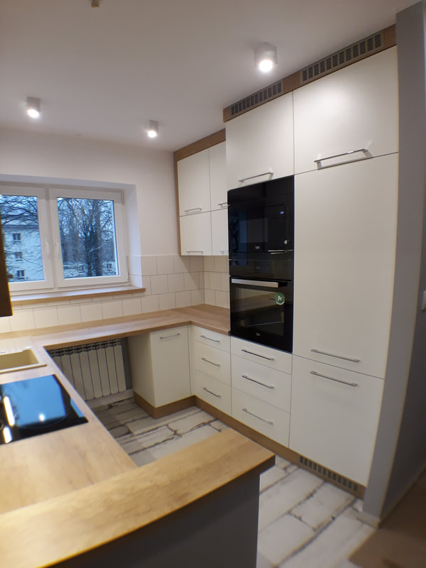 Jasna kuchnia na wymiar z drewnianymi blatami i zabudowaną czarną mikrofalówką oraz piekarnikiem, widok na okno z grzejnikiem.