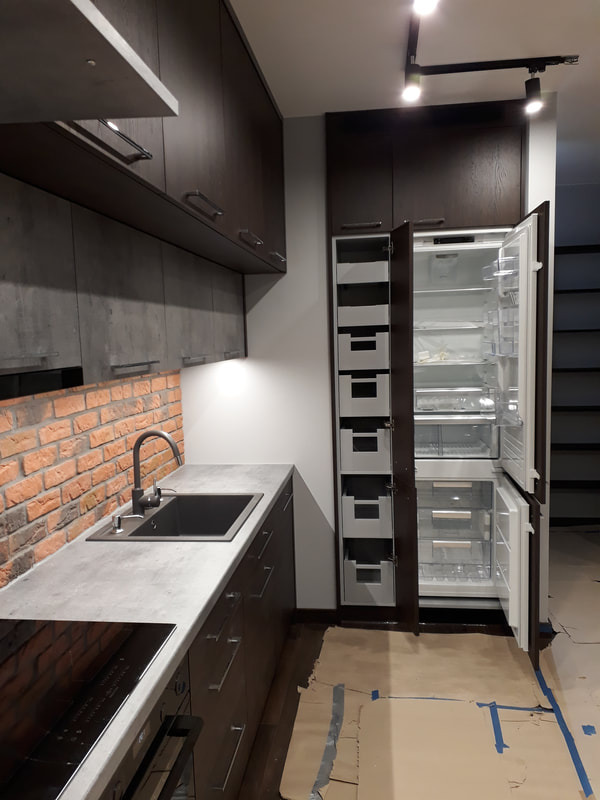 Nowoczesna kuchnia z ciemnymi, matowymi szafkami, blatem w odcieniach szarości i ceglaną ścianą, z widoczną otwartą lodówką z wewnętrznymi szufladami.
