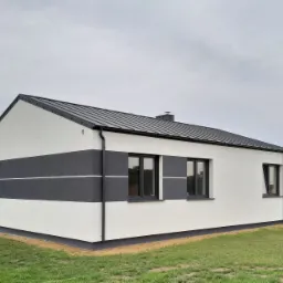 Nowoczesny dom jednorodzinny z białą elewacją i szarymi pasami, czarne okna i dach, widok z zewnątrz na tle zielonego trawnika i pochmurnego nieba.