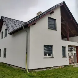 Dom jednorodzinny z szarą dachówką i jasną elewacją po tynkowaniu, widoczne rolety zewnętrzne, rynna i elementy konstrukcyjne dachu z drewna, teren wokół domu z zieloną trawą.