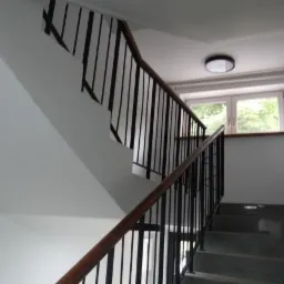 Klatka schodowa w bloku mieszkalnym z szarymi schodami, czarną balustradą z drewnianym poręczą i białymi ścianami, oświetlona lampą sufitową i naturalnym światłem z okna.