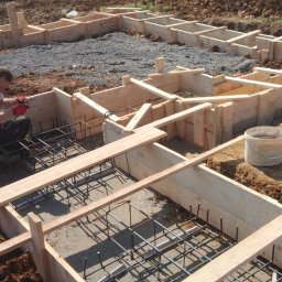 Eugeniusz Dobrovolskyi - Zbrojenie fundamentów z drewnianym szalunkiem na placu budowy z widocznym pracownikiem w czerwonych rękawicach i górą ziemi w tle.