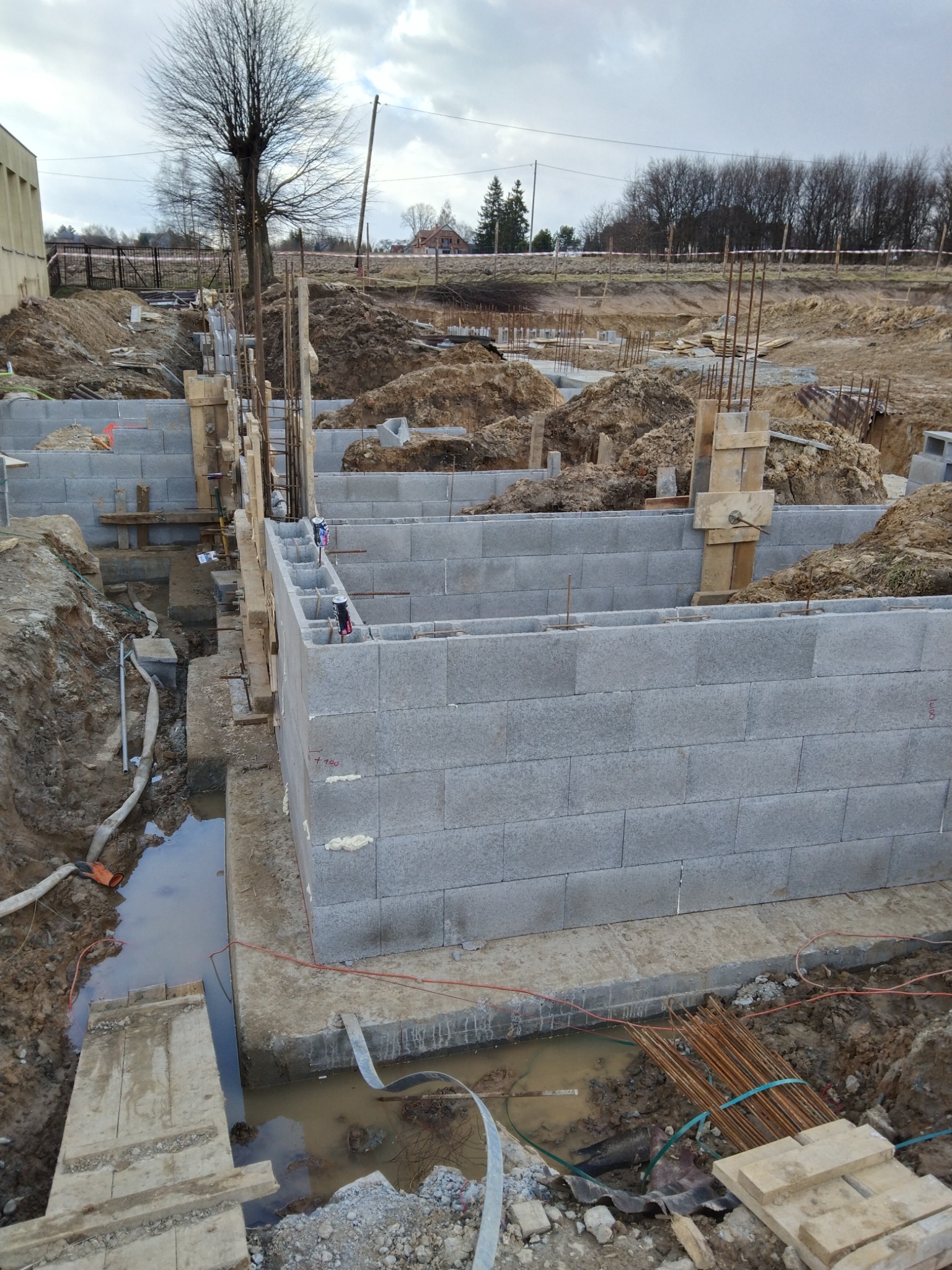 Wznoszenie fundamentów z bloczków betonowych na placu budowy, widoczne wystające pręty zbrojeniowe, drewniane szalunki i wykop z wodą.
