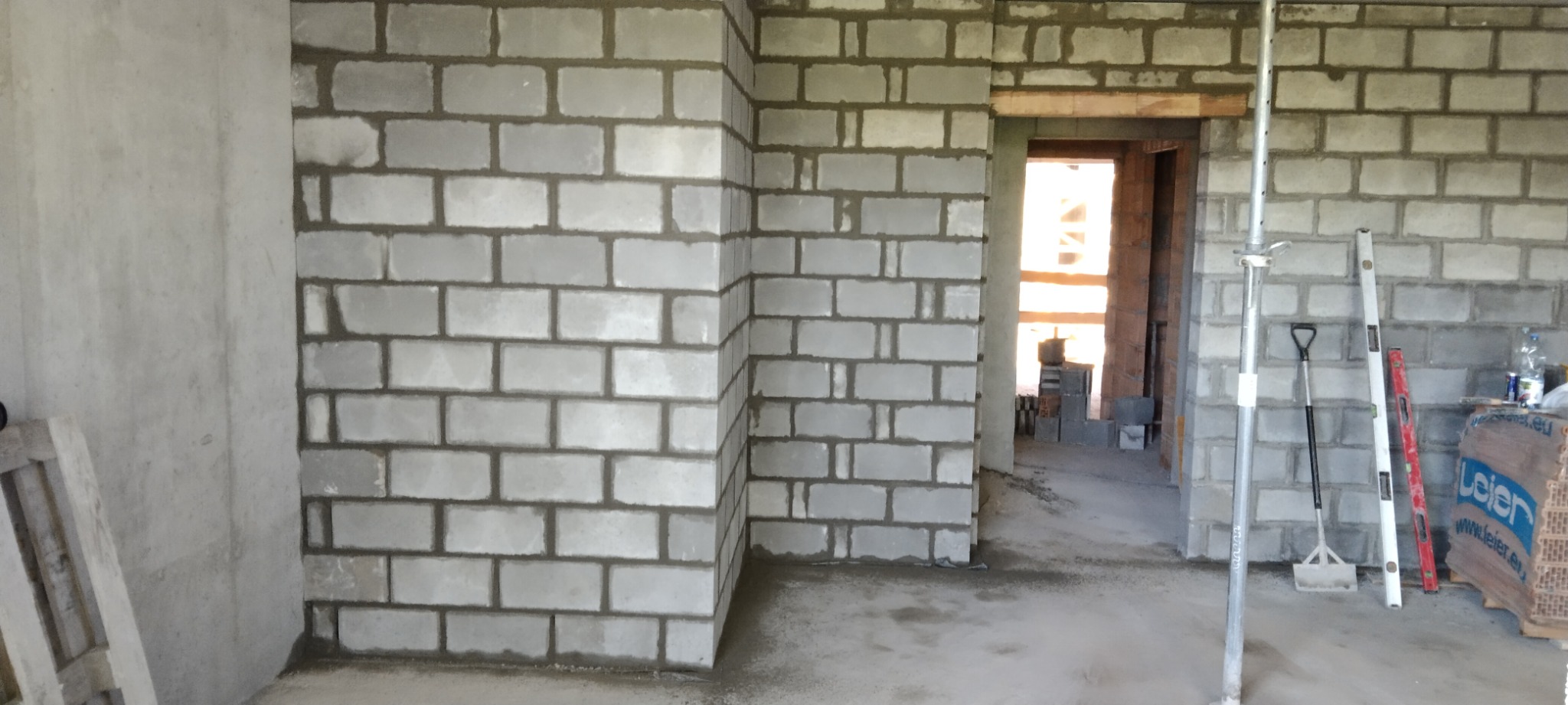 Wnętrze surowego domu w trakcie murowania ścian z betonu komórkowego, widoczne podpory stropowe, łopata i poziomica.