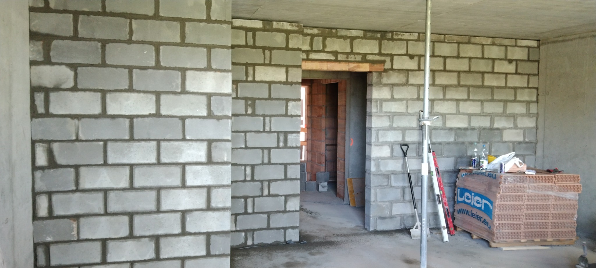 Wnętrze w trakcie budowy z widocznymi ścianami z szarych bloczków, otworami drzwiowymi, podporą stalową, narzędziami i paletą cegieł Leier w rogu pomieszczenia.