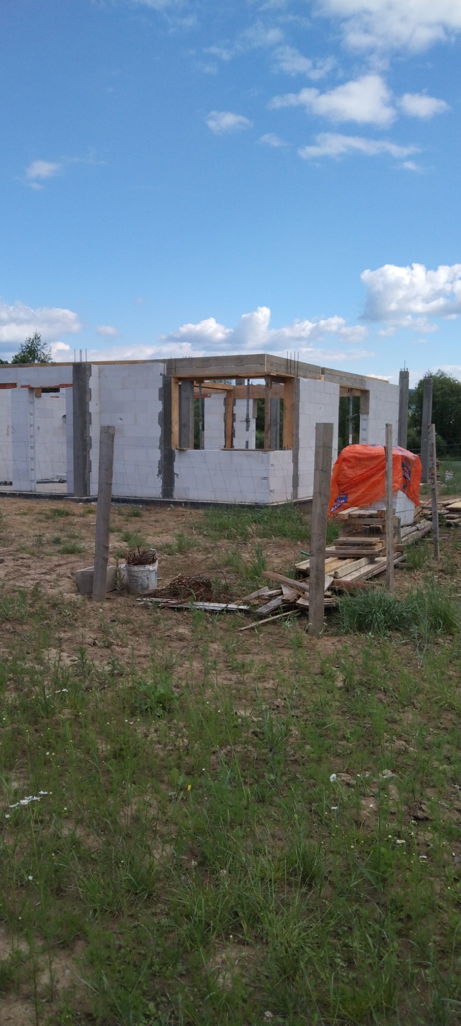 Wznoszona konstrukcja domu z betonu komórkowego, widoczne drewniane nadproża i zbrojenia, na pierwszym planie składowane materiały budowlane przykryte pomarańczową folią.