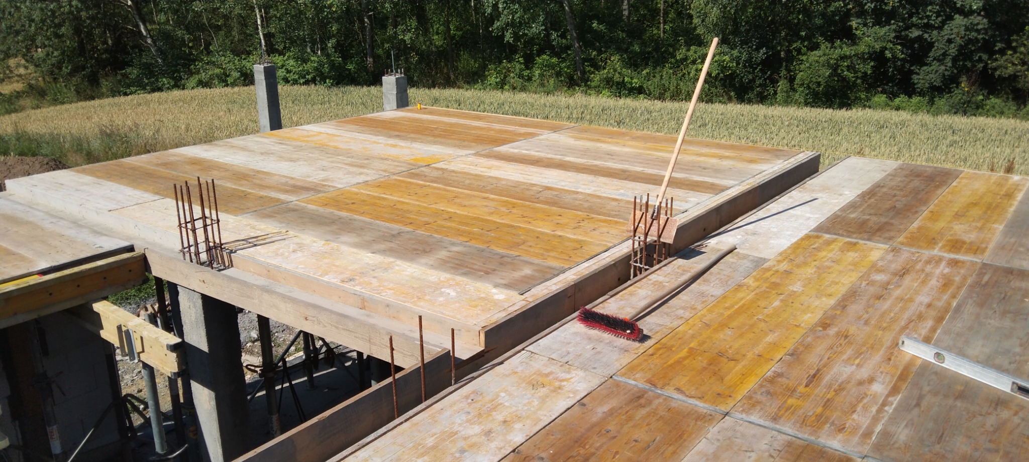 Zalany fundament z wystającymi prętami zbrojeniowymi, szalunek z desek, widok z góry na tle zieleni i pola.