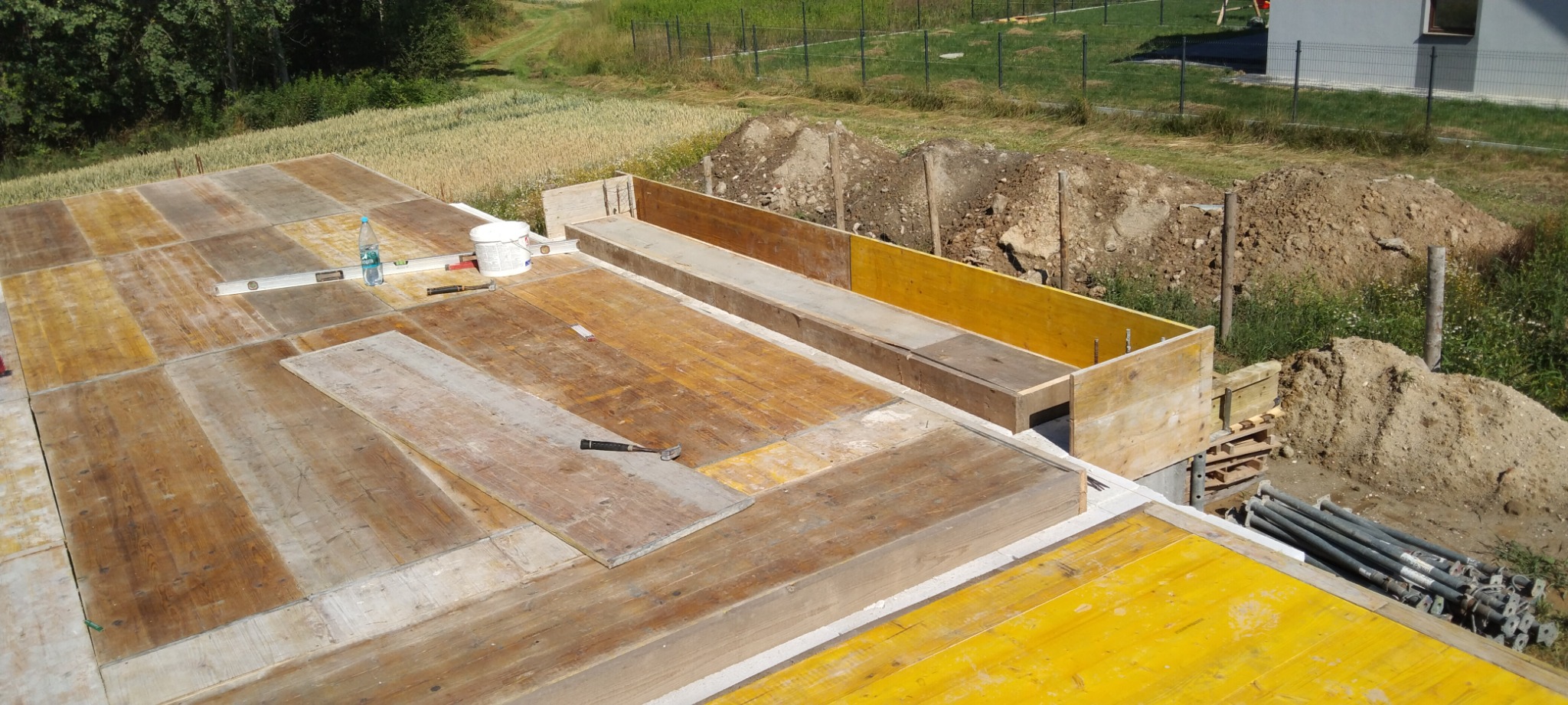 Przygotowanie szalunku pod fundamenty domu jednorodzinnego, widoczne deski szalunkowe, poziomica, młotek i wiadro z zaprawą, w tle hałda ziemi.