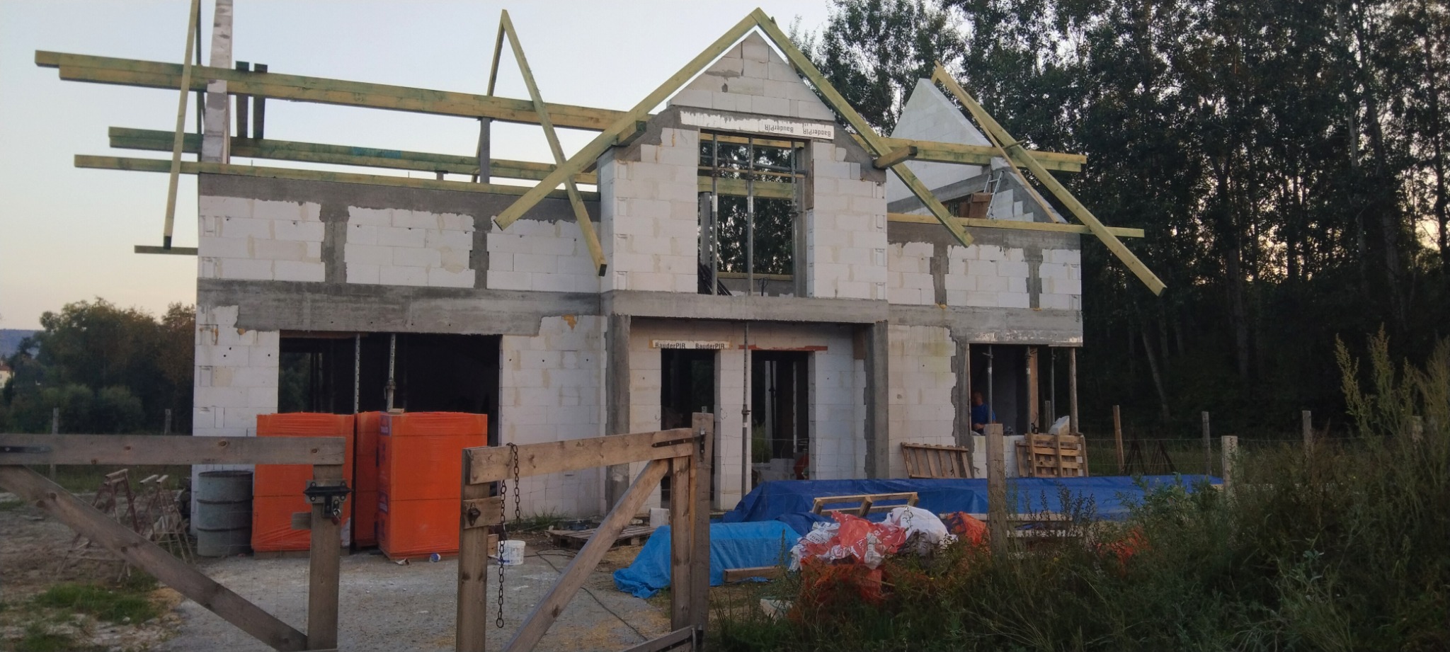 Surowy stan dwukondygnacyjnego domu z białych bloczków, z widoczną konstrukcją dachu z drewnianych belek i pustymi otworami okiennymi, na pierwszym planie pomarańczowe paczki materiałów budowlanych.