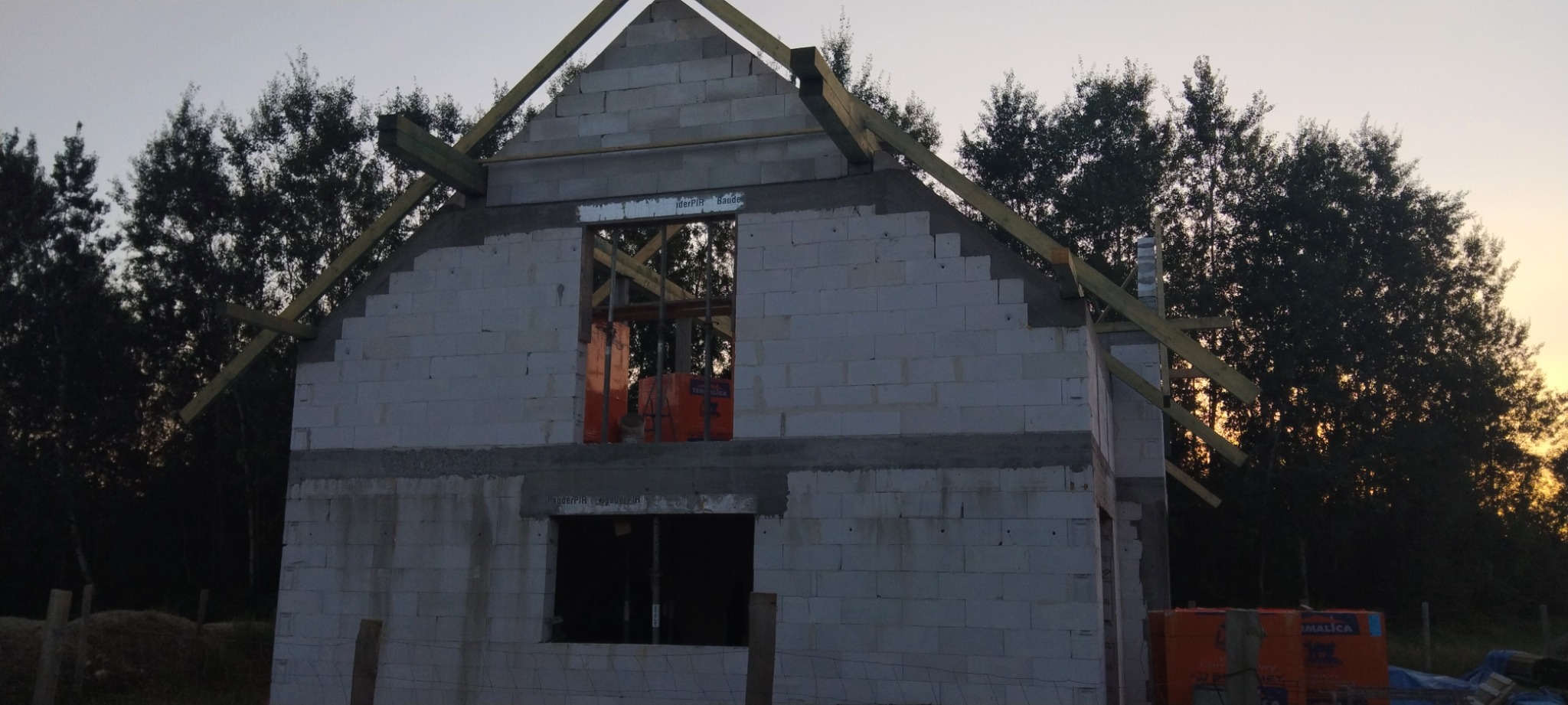 Wznoszenie ścian domu z bloczków w Krakowie, widoczne drewniane elementy konstrukcji dachu i paleta z materiałami budowlanymi.