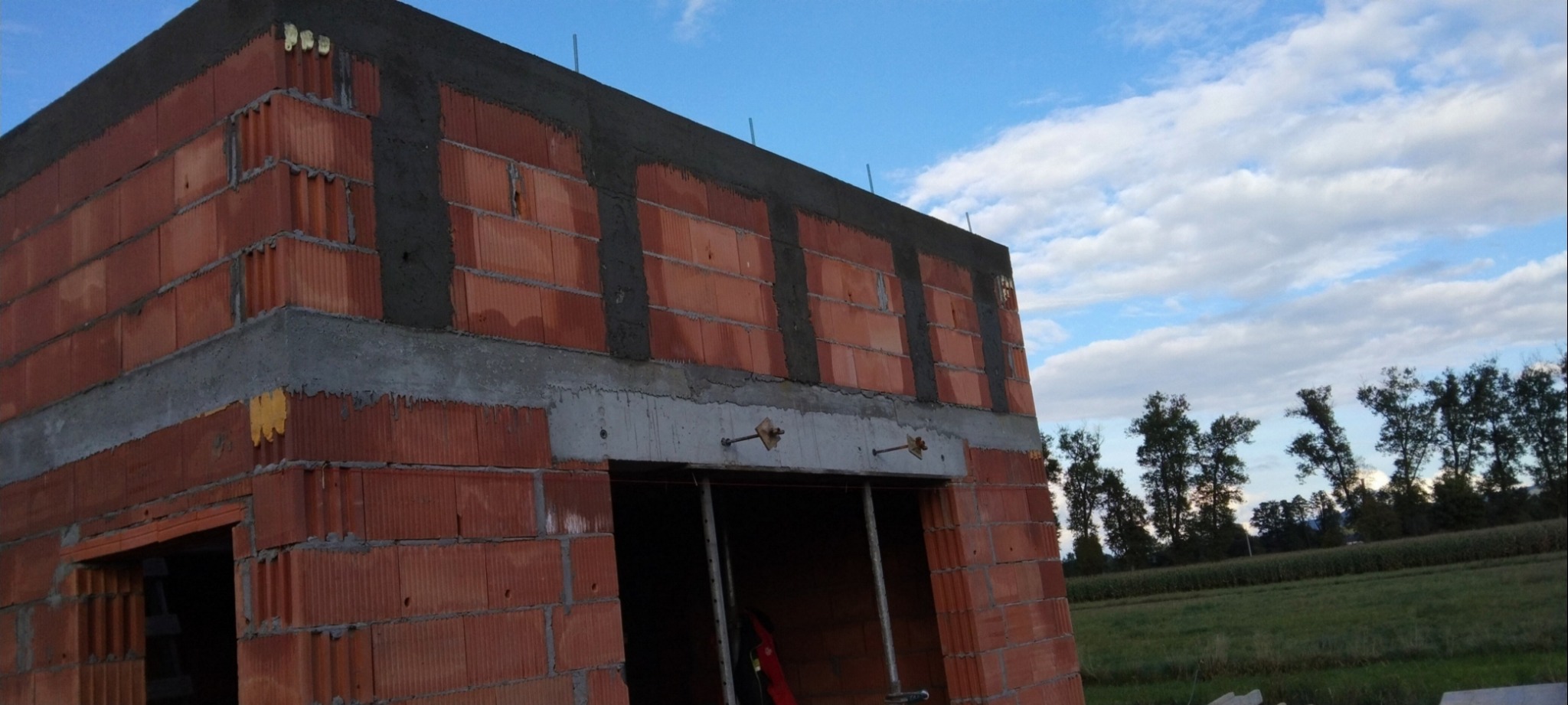 Wznoszenie ścian z czerwonej cegły ceramicznej, widoczne świeże spoiny i zbrojenia, w tle błękitne niebo i zieleń drzew.