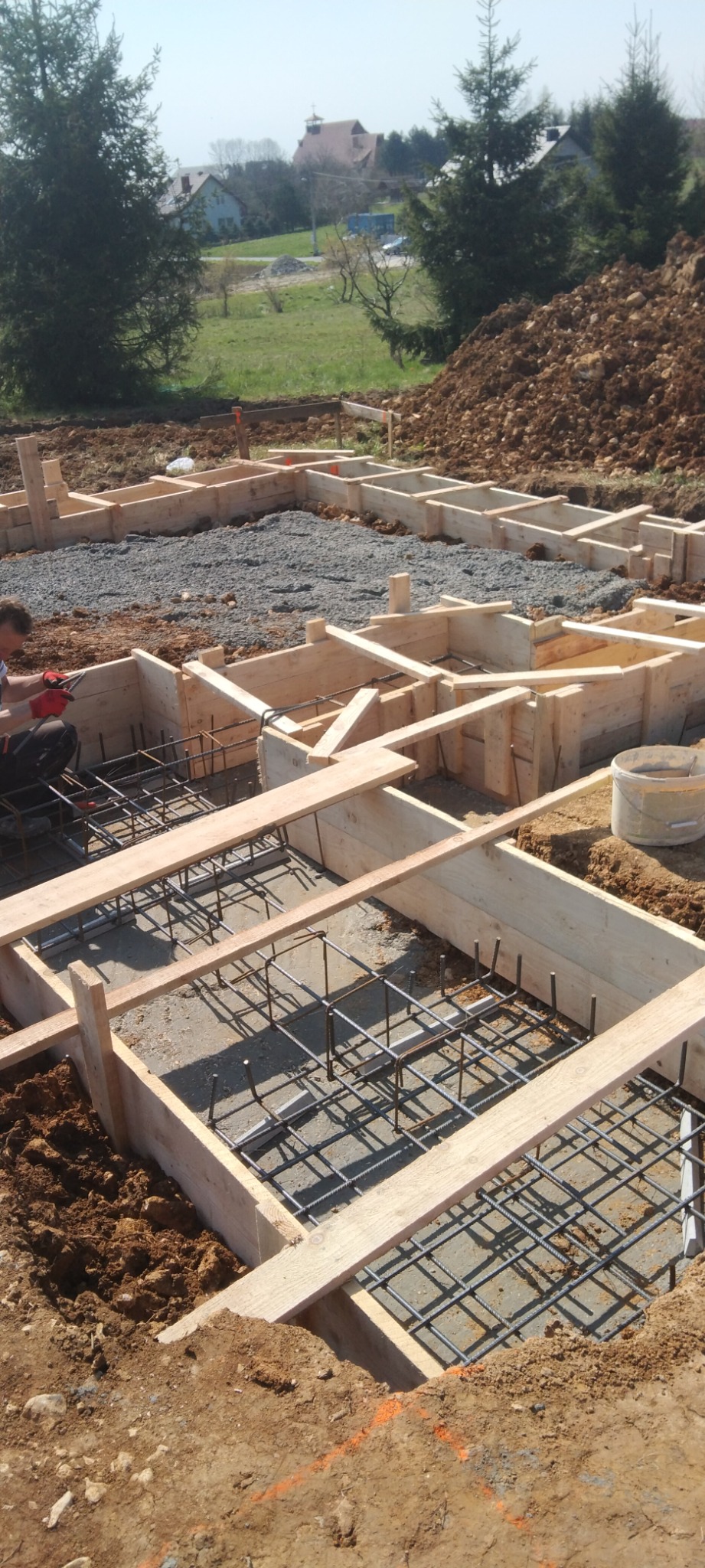 Zbrojenie fundamentów z drewnianym szalunkiem na placu budowy z widocznym pracownikiem w czerwonych rękawicach i górą ziemi w tle.