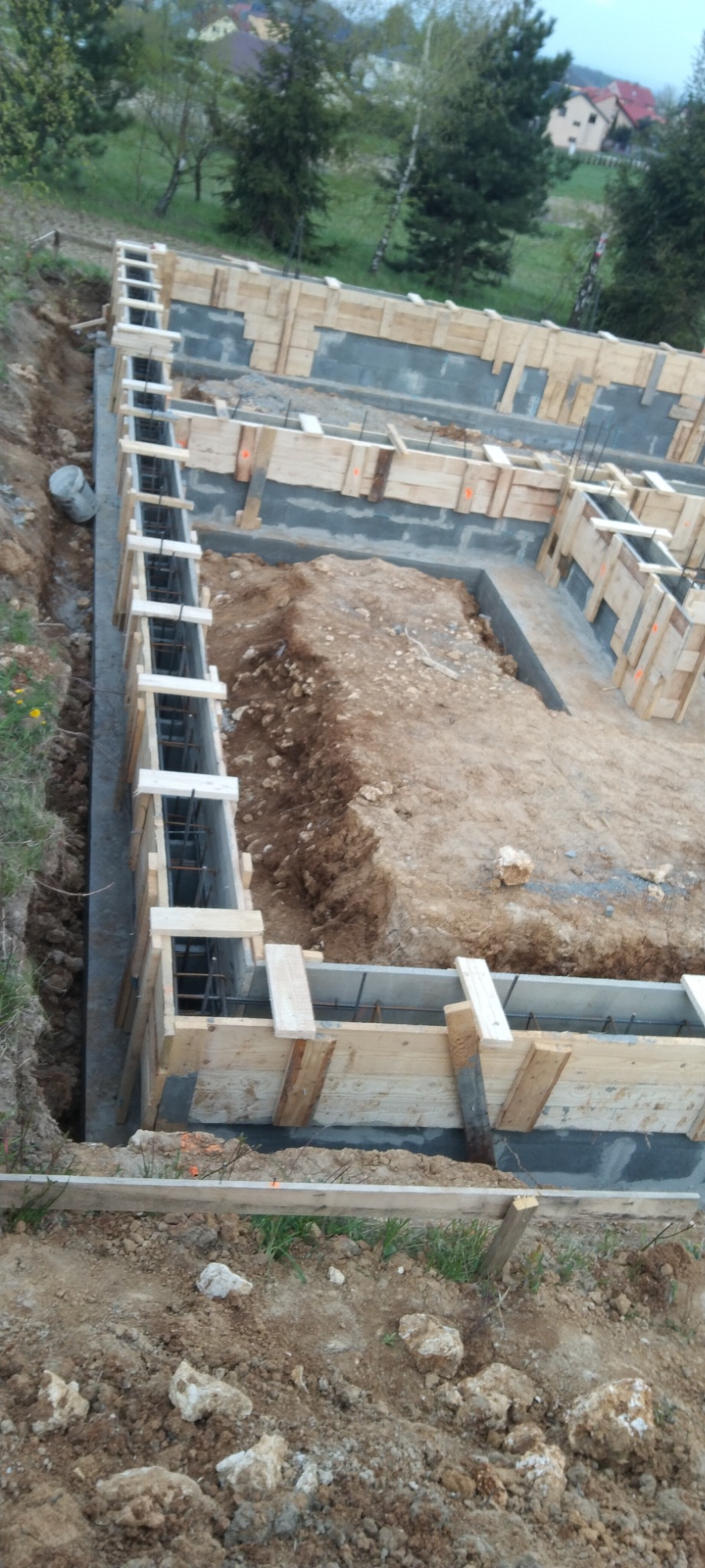 Wykopane fundamenty z betonową wylewką i drewnianym szalunkiem wzmocnione stalowymi prętami, widok z góry na tle zieleni i zabudowań.