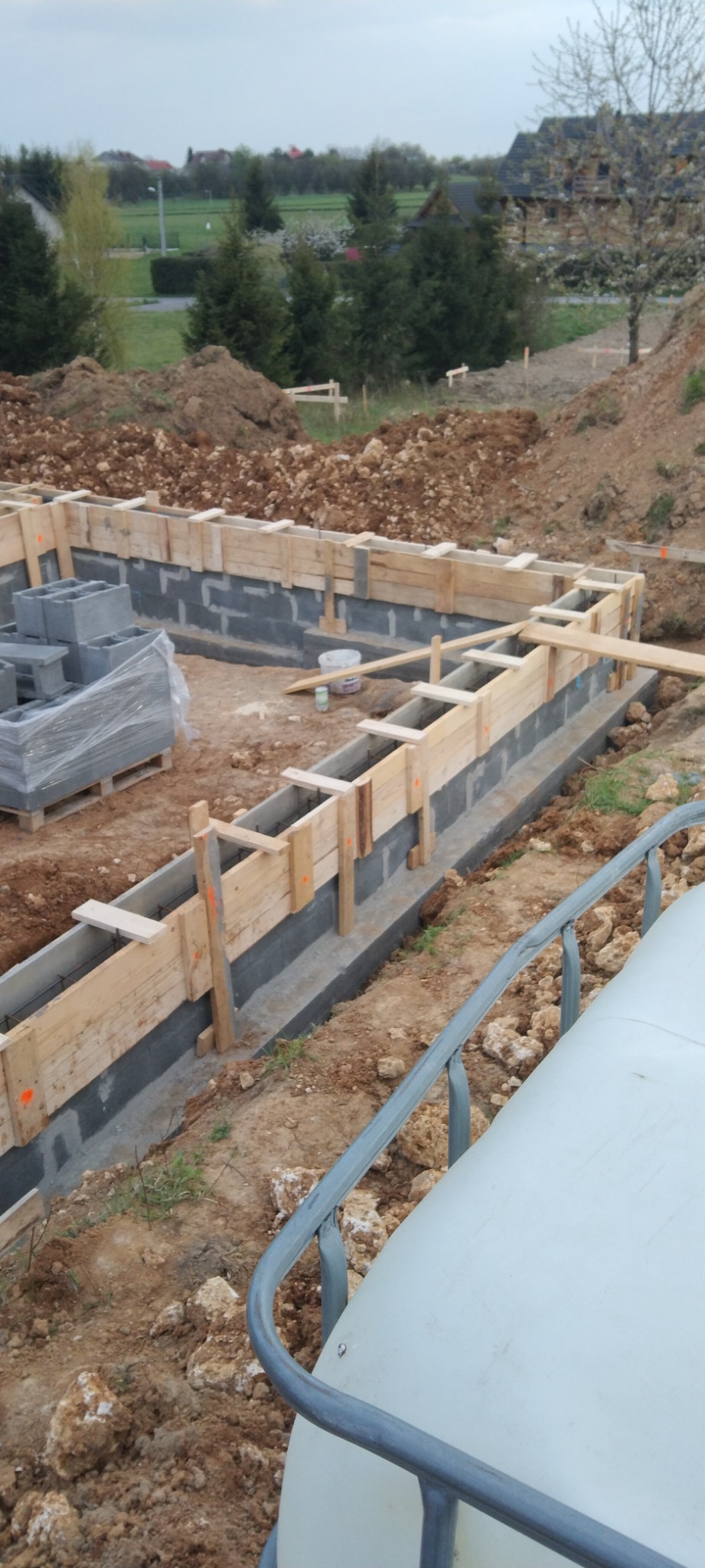 Przygotowanie fundamentów pod budowę domu: szalunek z desek, zbrojenie i częściowo wylany beton. W tle widoczny teren budowy i zabudowania.