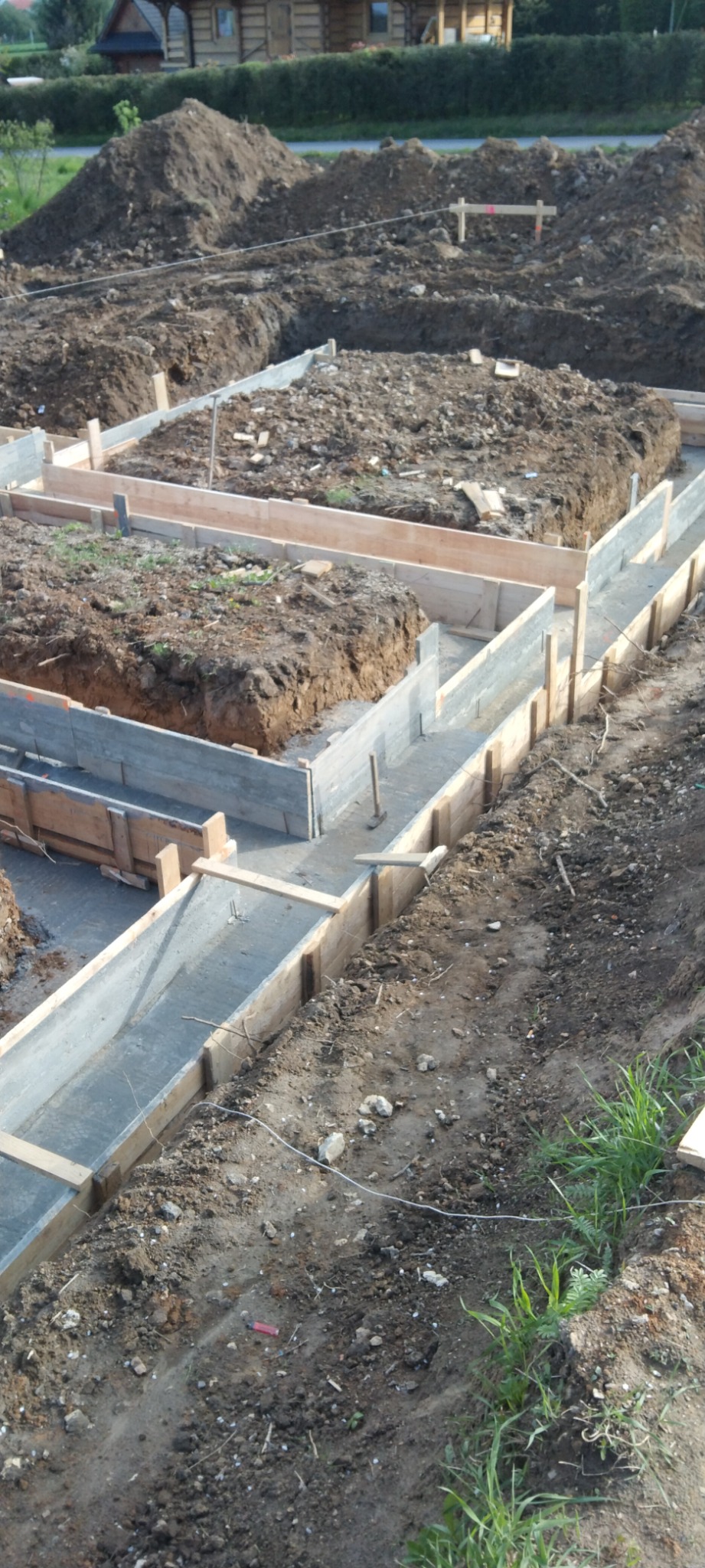 Wykop pod fundamenty z deskowaniem i częściowo wylanym betonem, widoczne narzędzia i ziemia, w tle drewniany dom.