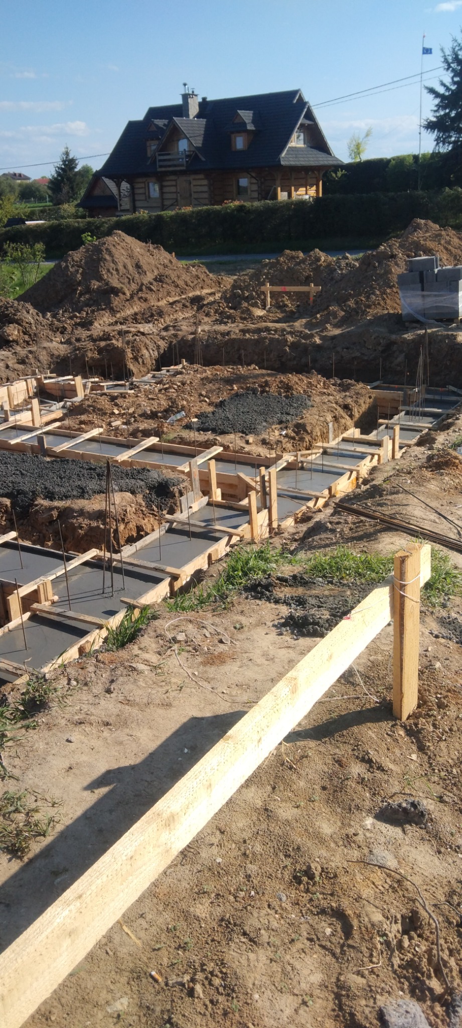 Wykopane fundamenty pod dom z drewnianą konstrukcją szkieletową w tle, częściowo zalane betonem, z widocznym zbrojeniem i szalunkami.