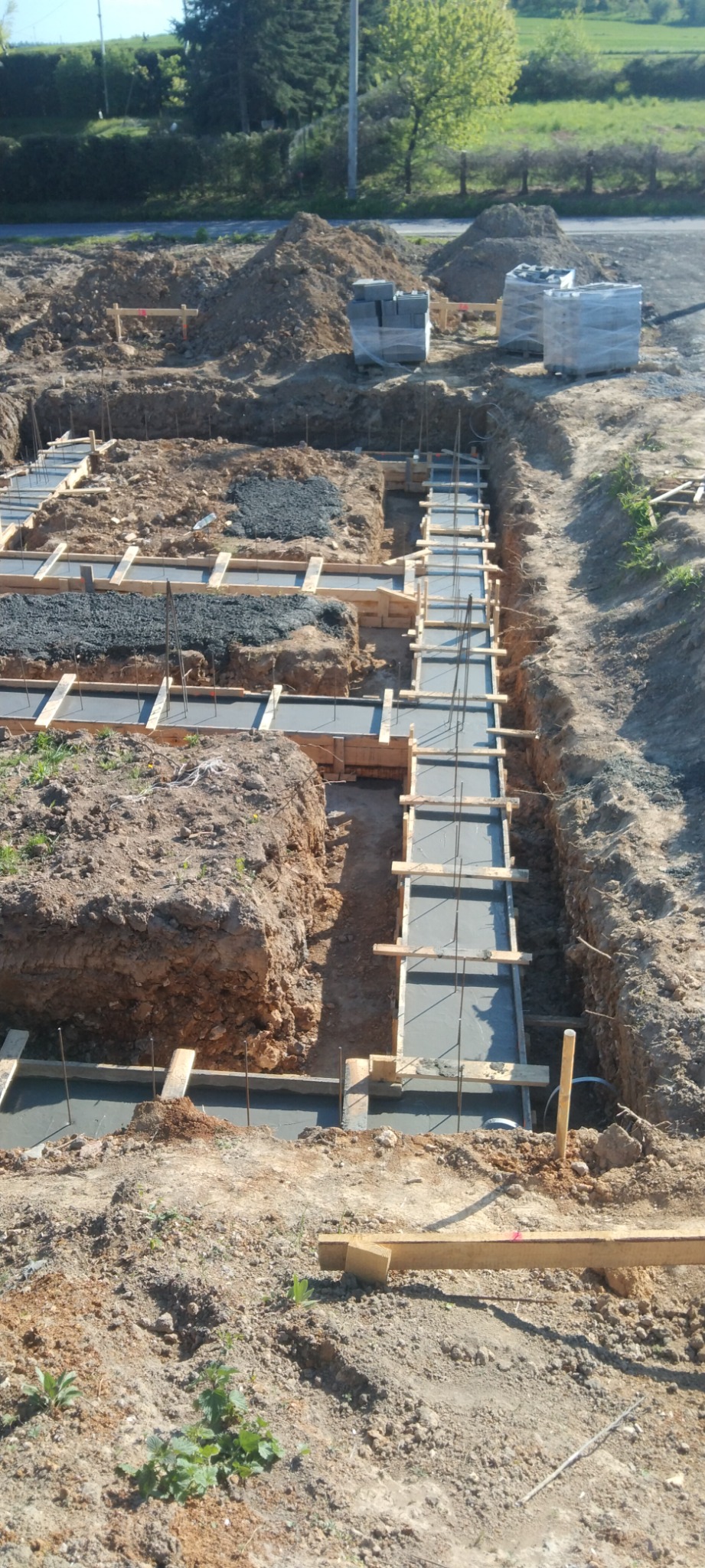 Wykopane fundamenty z drewnianym szalunkiem i zalanym betonem, widoczne zbrojenie, w tle ułożone bloczki fundamentowe.