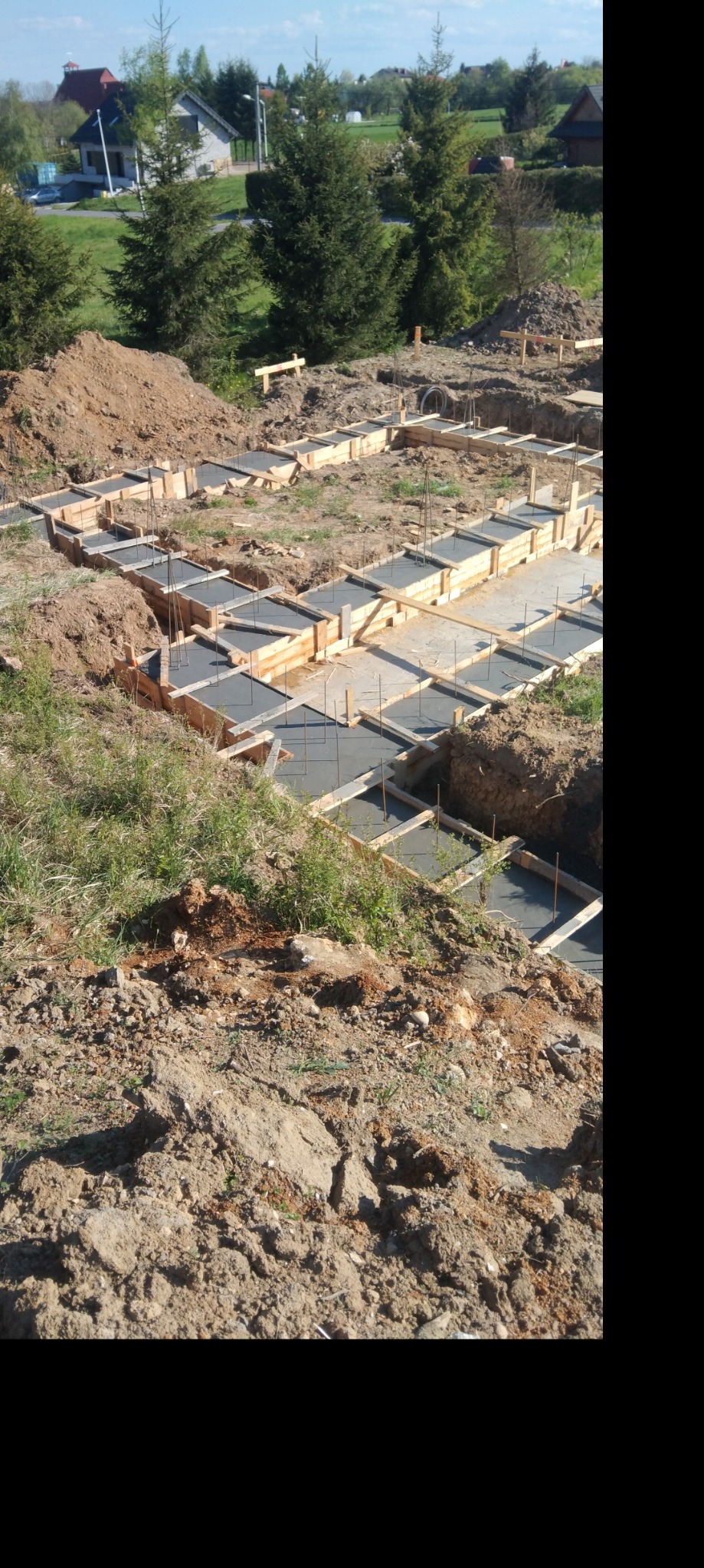 Wylane fundamenty z drewnianym szalunkiem i wystającymi prętami zbrojeniowymi na tle zieleni i zabudowań.