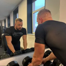 Mężczyzna w czarnej koszulce z logo Supermana, przygotowuje się do podnoszenia hantli przed lustrem w siłowni.