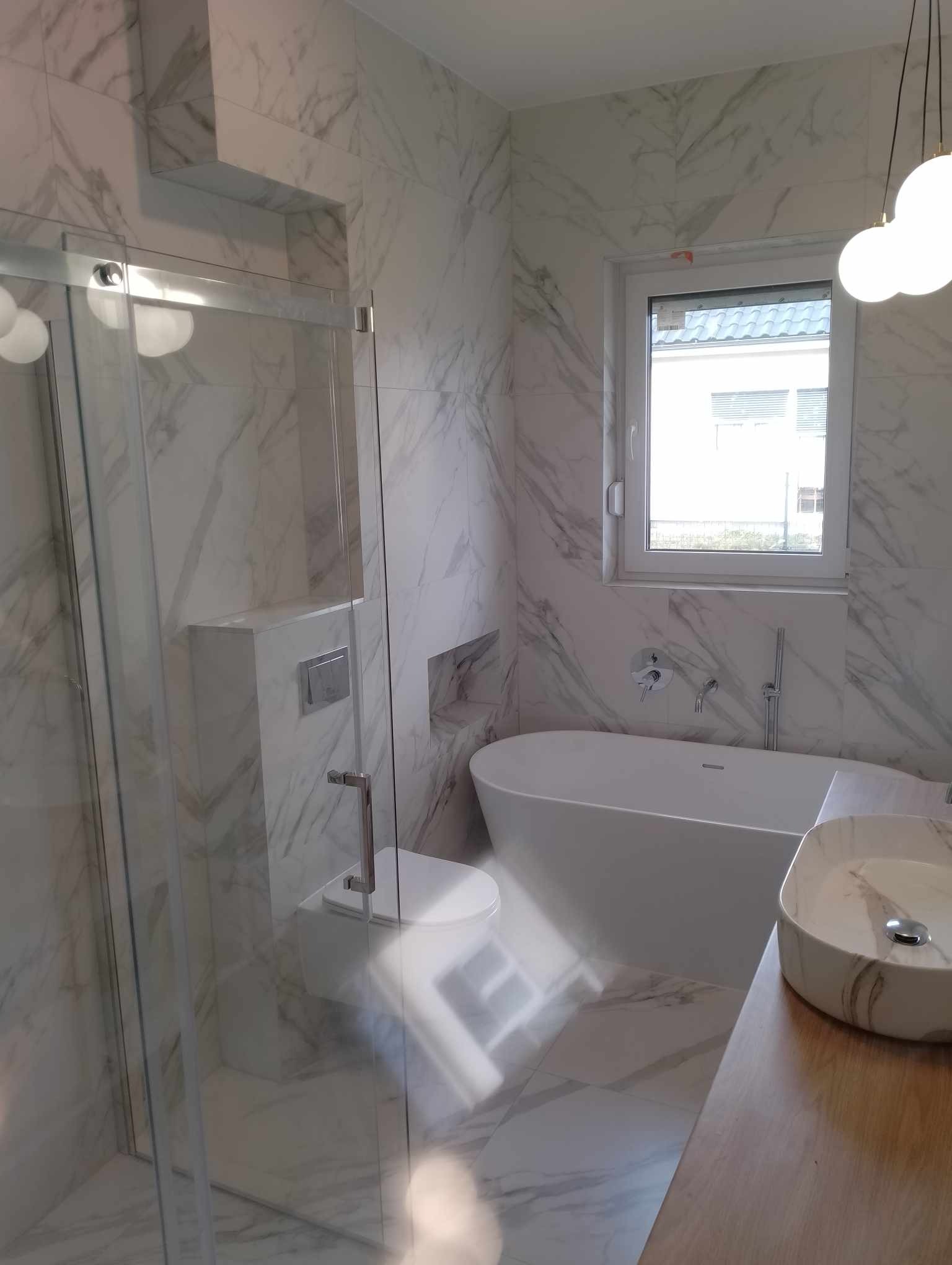 Nowoczesna łazienka z marmurowymi płytkami, kabiną prysznicową ze szklanymi drzwiami przesuwnymi, wolnostojącą wanną przy oknie, umywalką nablatową i podwieszaną toaletą.