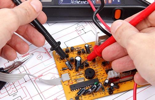 Pomiar napięcia na płycie elektronicznej alarmu za pomocą multimetru, widoczne schematy elektryczne oraz szczypce