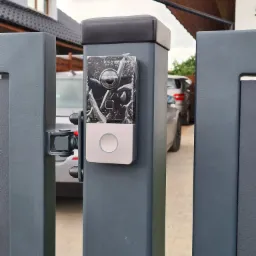 Nowy wideodomofon z kamerą Hikvision, jeszcze w folii ochronnej, zamontowany na szarym słupku ogrodzenia posesji. W tle widoczny fragment domu i samochód.