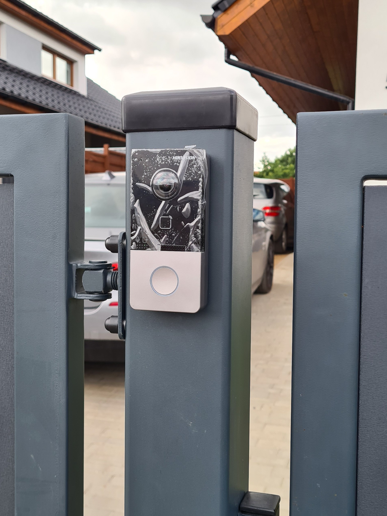 Nowy wideodomofon z kamerą Hikvision, jeszcze w folii ochronnej, zamontowany na szarym słupku ogrodzenia posesji. W tle widoczny fragment domu i samochód.