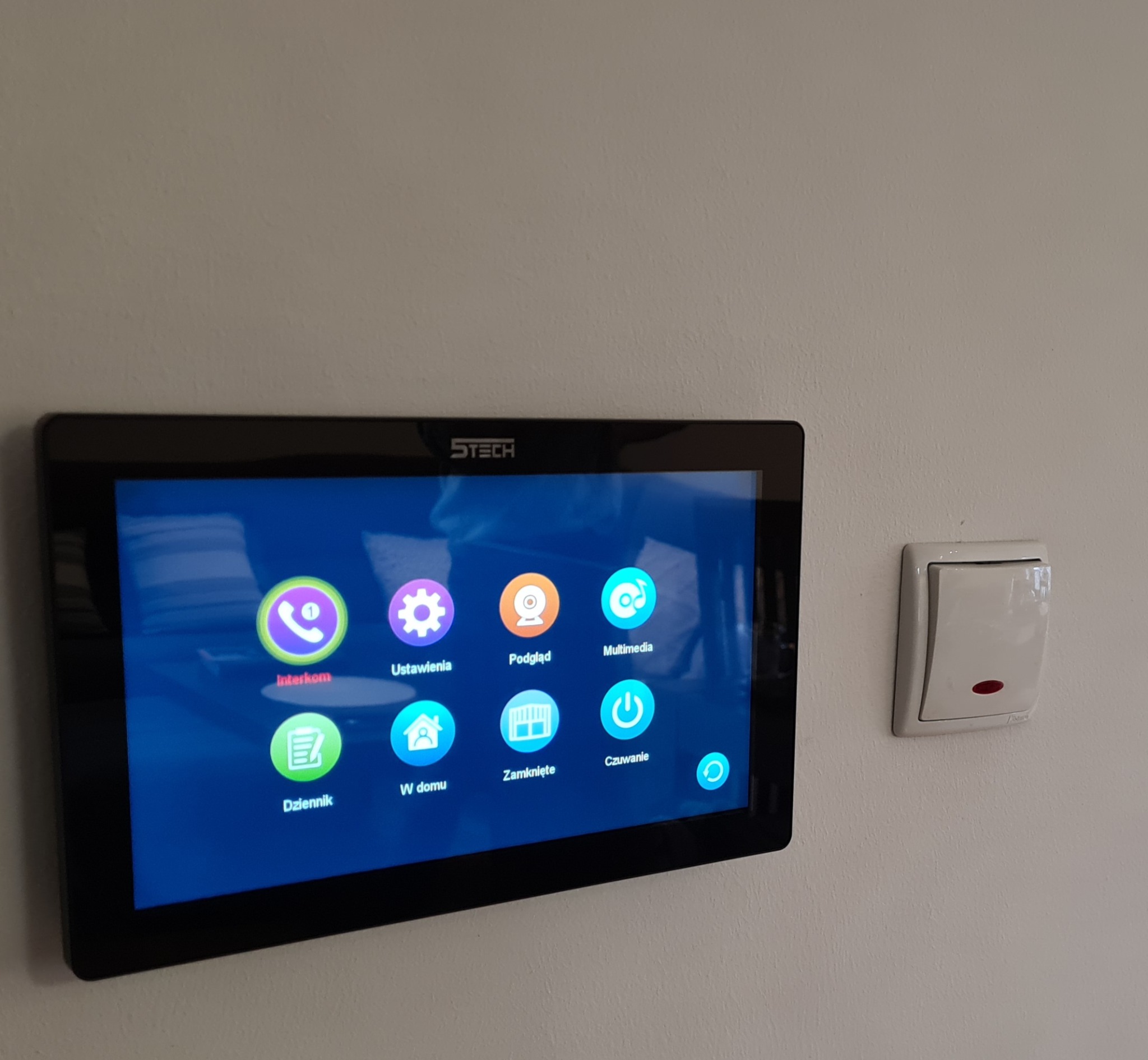 Panel dotykowy wideodomofonu marki STech zamontowany na ścianie, widoczne ikony interfejsu użytkownika: Interkom, Ustawienia, Podgląd, Multimedia, Dziennik, W domu, Zamknięte, Czuwanie. Obok...