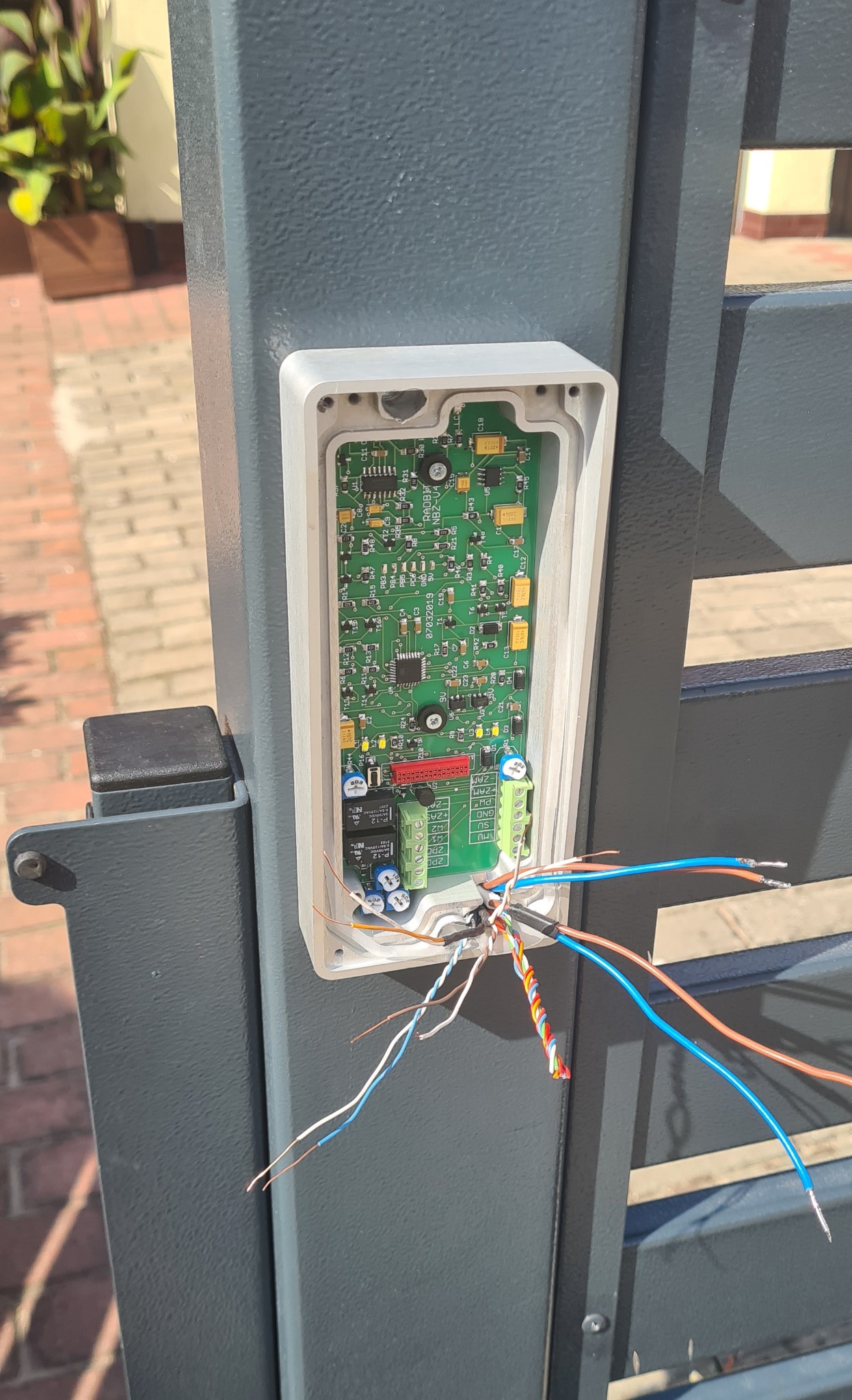 Otwarta obudowa panelu sterującego systemu dostępu, widoczna płytka drukowana z elementami elektronicznymi i podłączone przewody z różnymi kolorami izolacji, zamontowana na szarym słupku bramy.