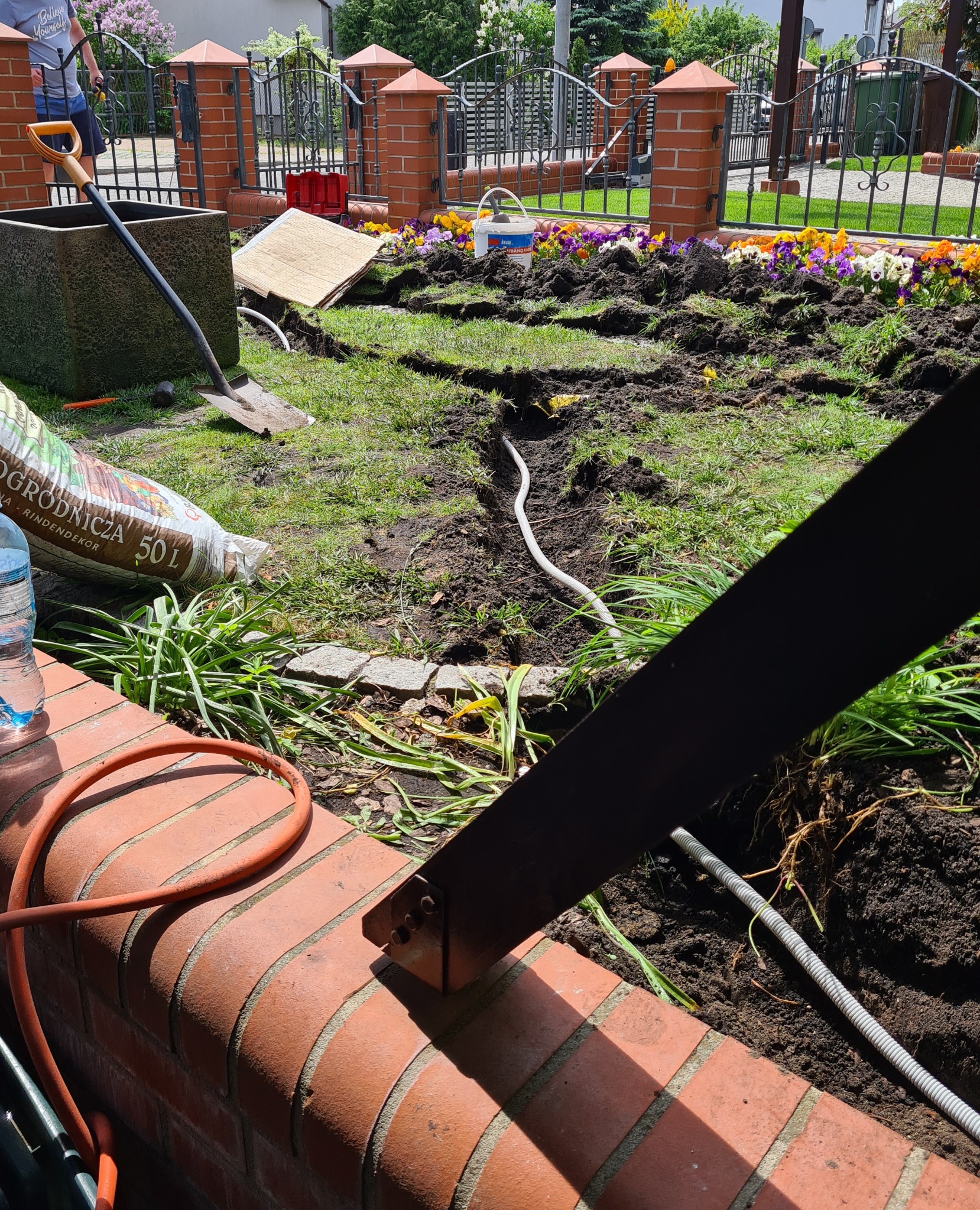 Wykonane rowy w ziemi pod instalację kabli, widoczne kwiaty, narzędzia ogrodnicze i worki z ziemią.