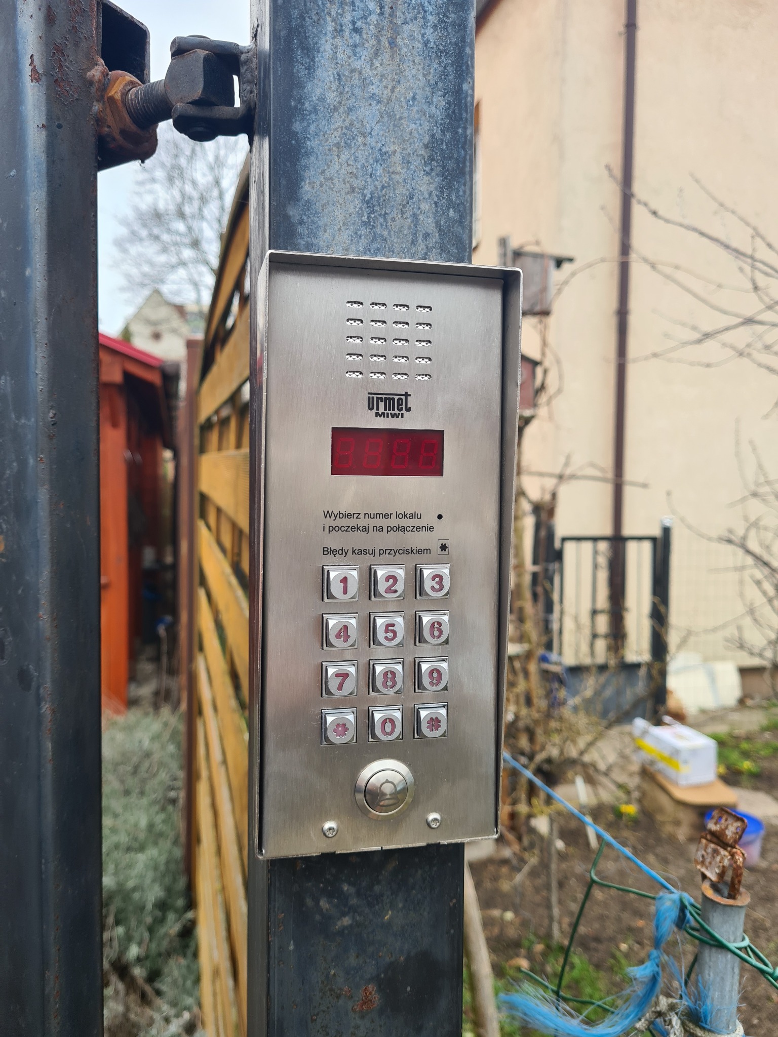 Domofon Urmet Miwi z klawiaturą numeryczną zamontowany na metalowej bramie, widoczne ślady korozji na zawiasach bramy, w tle budynki i zieleń.