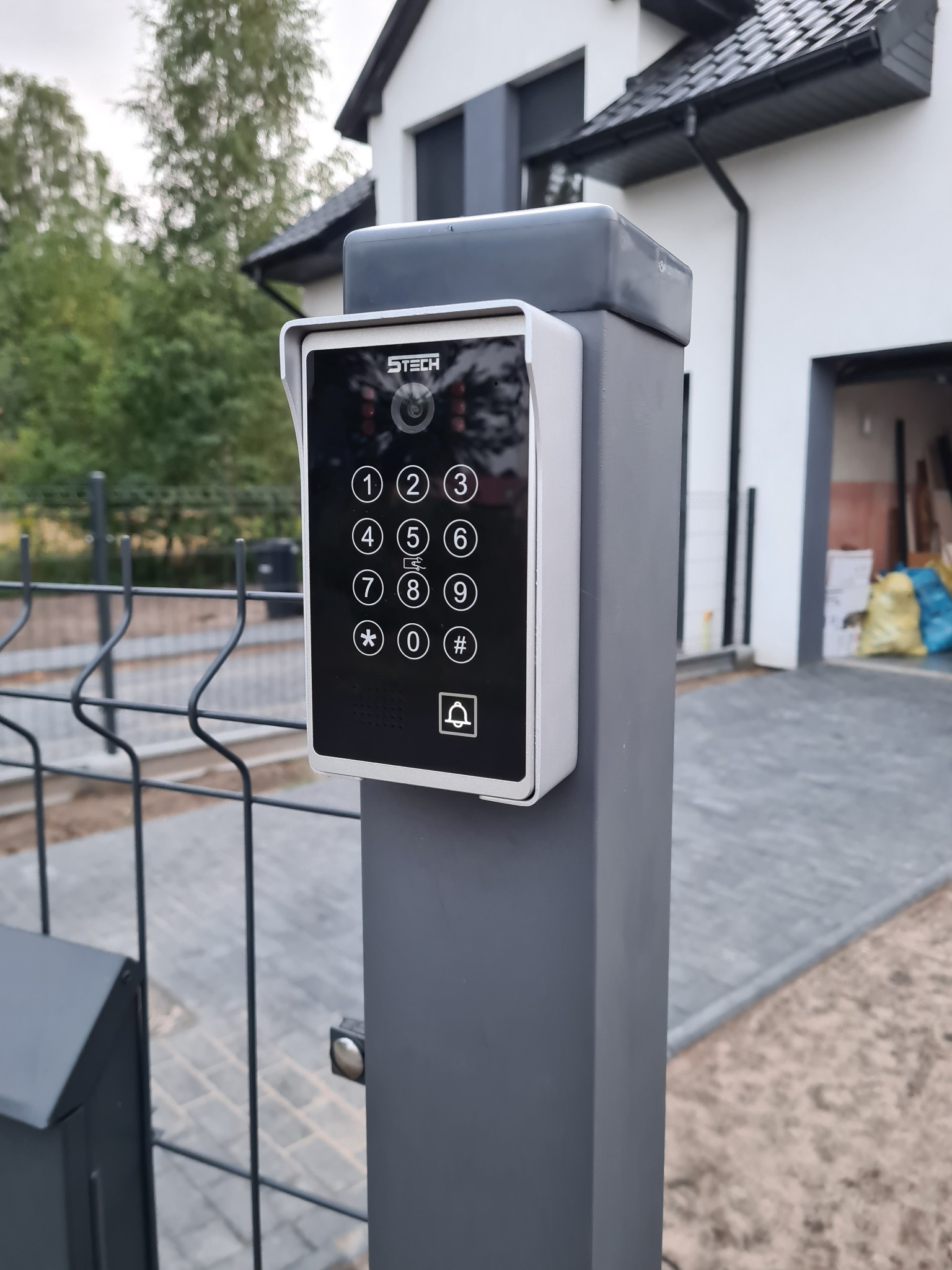 Zewnętrzny panel dotykowy domofonu z klawiaturą numeryczną i kamerą, zamontowany na metalowym słupku ogrodzenia przed nowoczesnym domem z garażem.