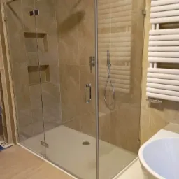 Nowoczesna kabina prysznicowa z transparentnego szkła, zamontowana w łazience wykończonej beżowymi płytkami imitującymi marmur, widoczny grzejnik drabinkowy i fragment wanny.