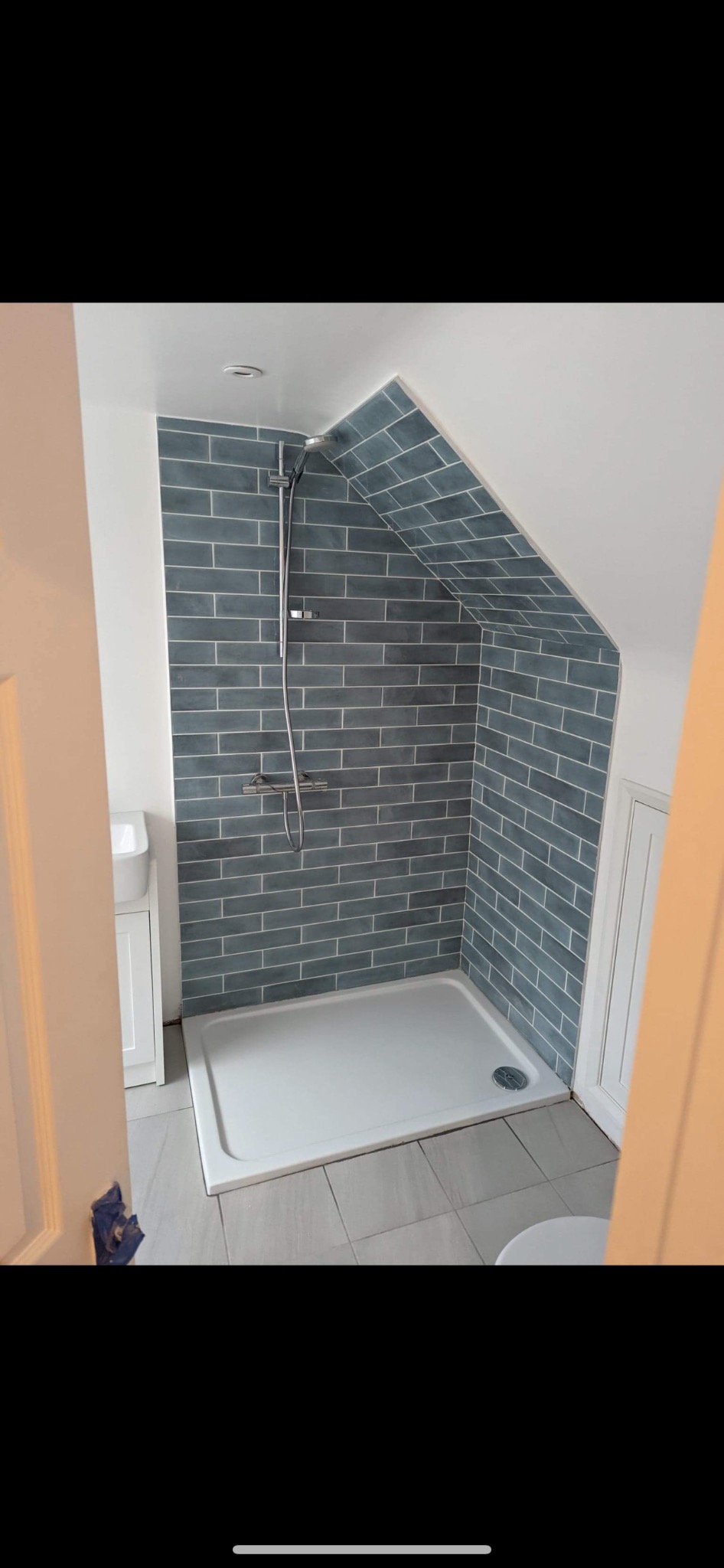 Nowoczesna kabina prysznicowa z szarymi, cegiełkowymi płytkami ułożonymi pod skosem, biały brodzik i armatura, wkomponowana w przestrzeń pod skosami dachu.