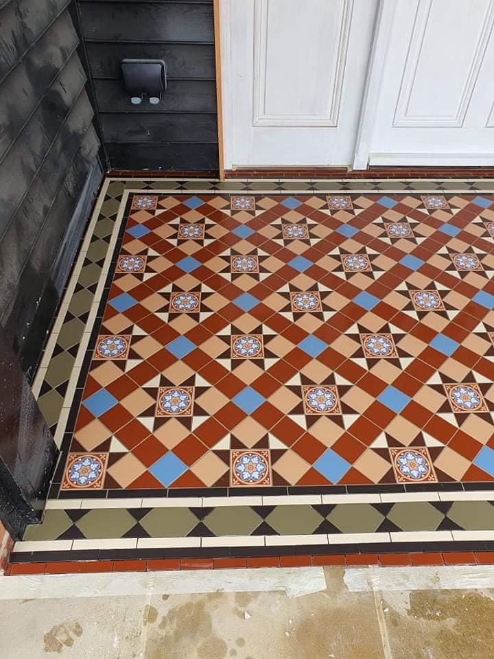 Wzorzysta, ceramiczna posadzka w holu wejściowym z motywem geometrycznym i kwiatowym, ułożona przy czarnych deskach elewacyjnych i białych drzwiach.