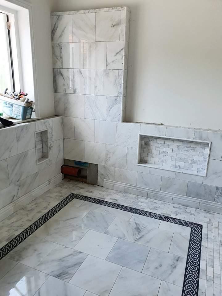 Łazienka w trakcie wykańczania z marmurowymi płytkami na ścianach i podłodze, widoczne wnęki w ścianach, jedna wykończona mozaiką, oraz czarna bordiura na podłodze.