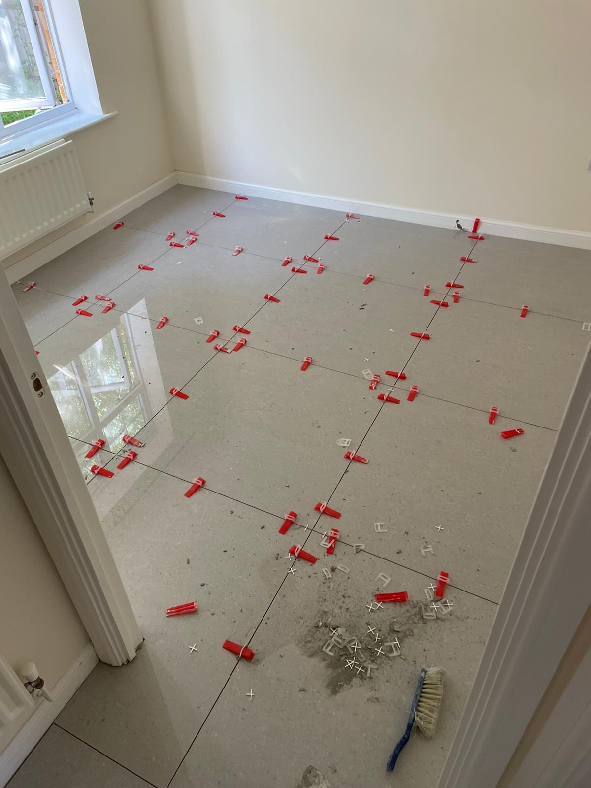 Szare płytki podłogowe z czerwonymi klipsami poziomującymi i krzyżykami dystansowymi podczas układania, widoczna szczotka do fugowania i pozostałości zaprawy.