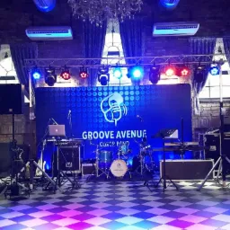Scena z nagłośnieniem i oświetleniem w ceglanym wnętrzu, gotowa na występ zespołu muzycznego Groove Avenue Cover Band, z widocznym perkusją, klawiszami i laptopem.