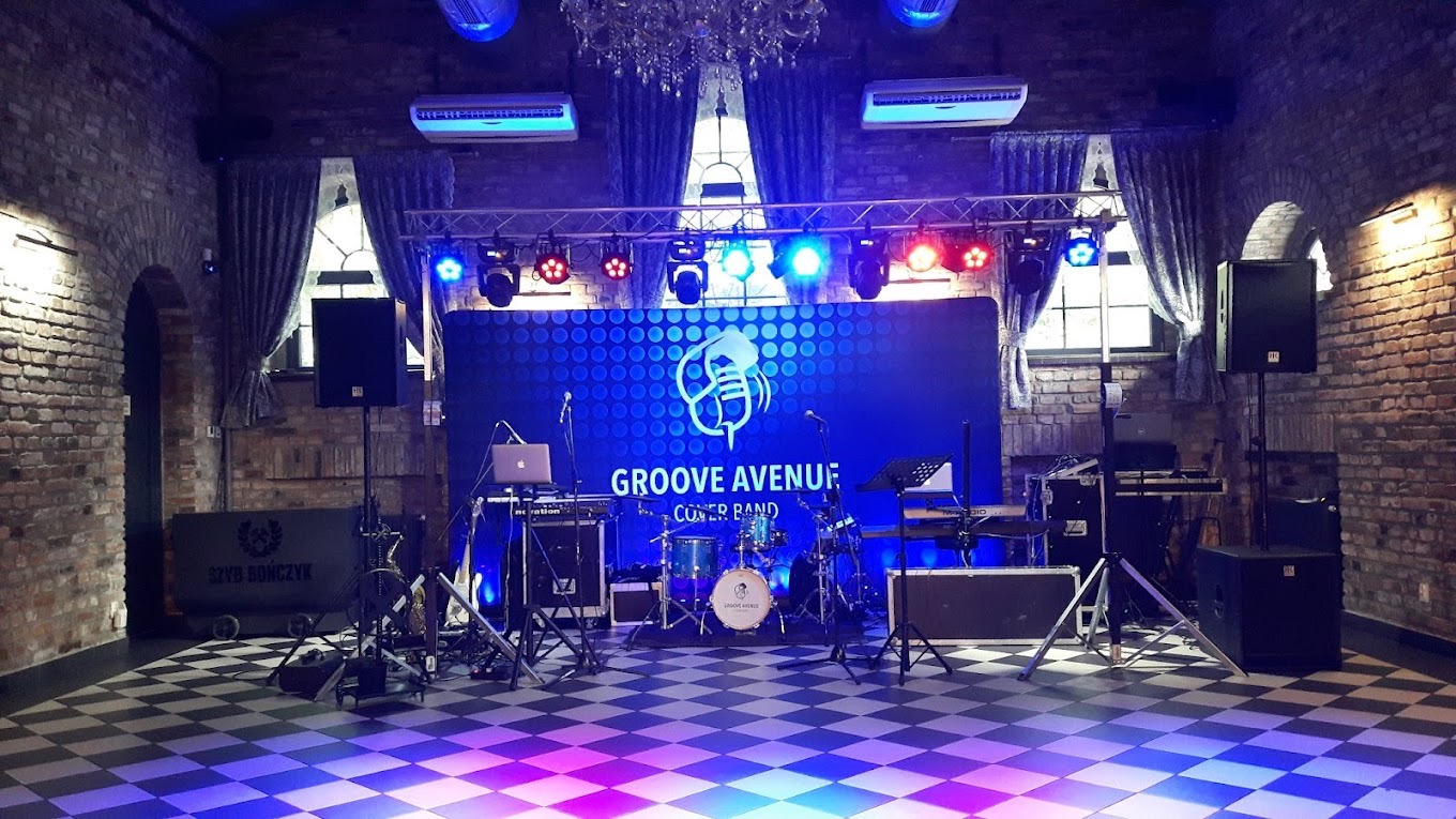 Scena z nagłośnieniem i oświetleniem w ceglanym wnętrzu, gotowa na występ zespołu muzycznego Groove Avenue Cover Band, z widocznym perkusją, klawiszami i laptopem.