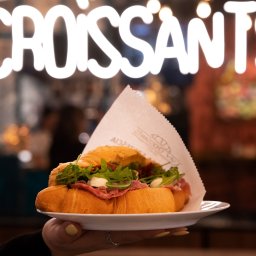 Dato Dariusz Grabiec - Kadr z bliska: ręka trzyma biały talerz z croissantem wypełnionym szynką, rukolą i mozzarellą na tle neonowego napisu 'CROISSANTS' i rozmytego wnętrza kawiarni.