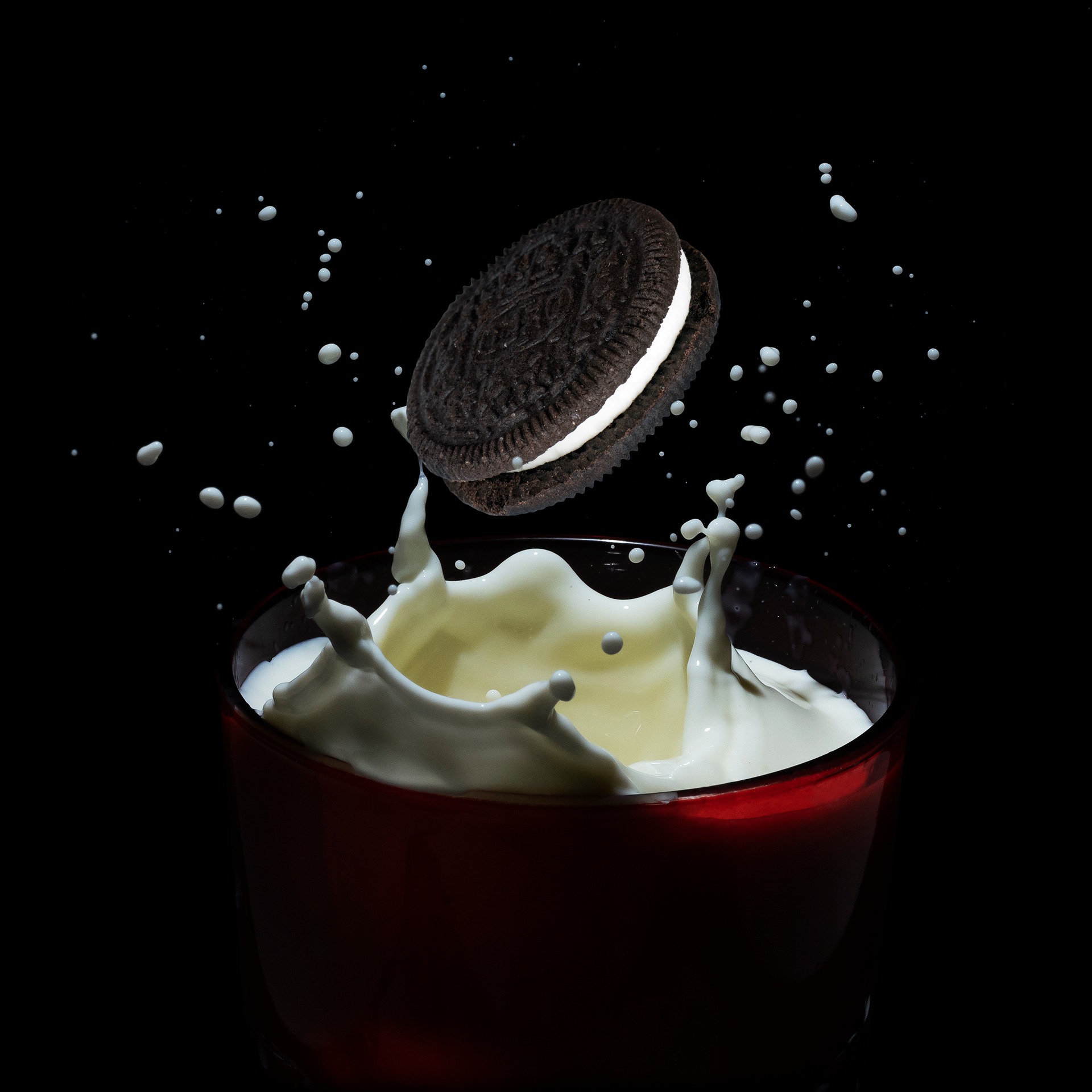Ciasteczko Oreo w locie nad szklanką mleka, z efektem rozprysku i kroplami zawieszonymi w powietrzu na czarnym tle.