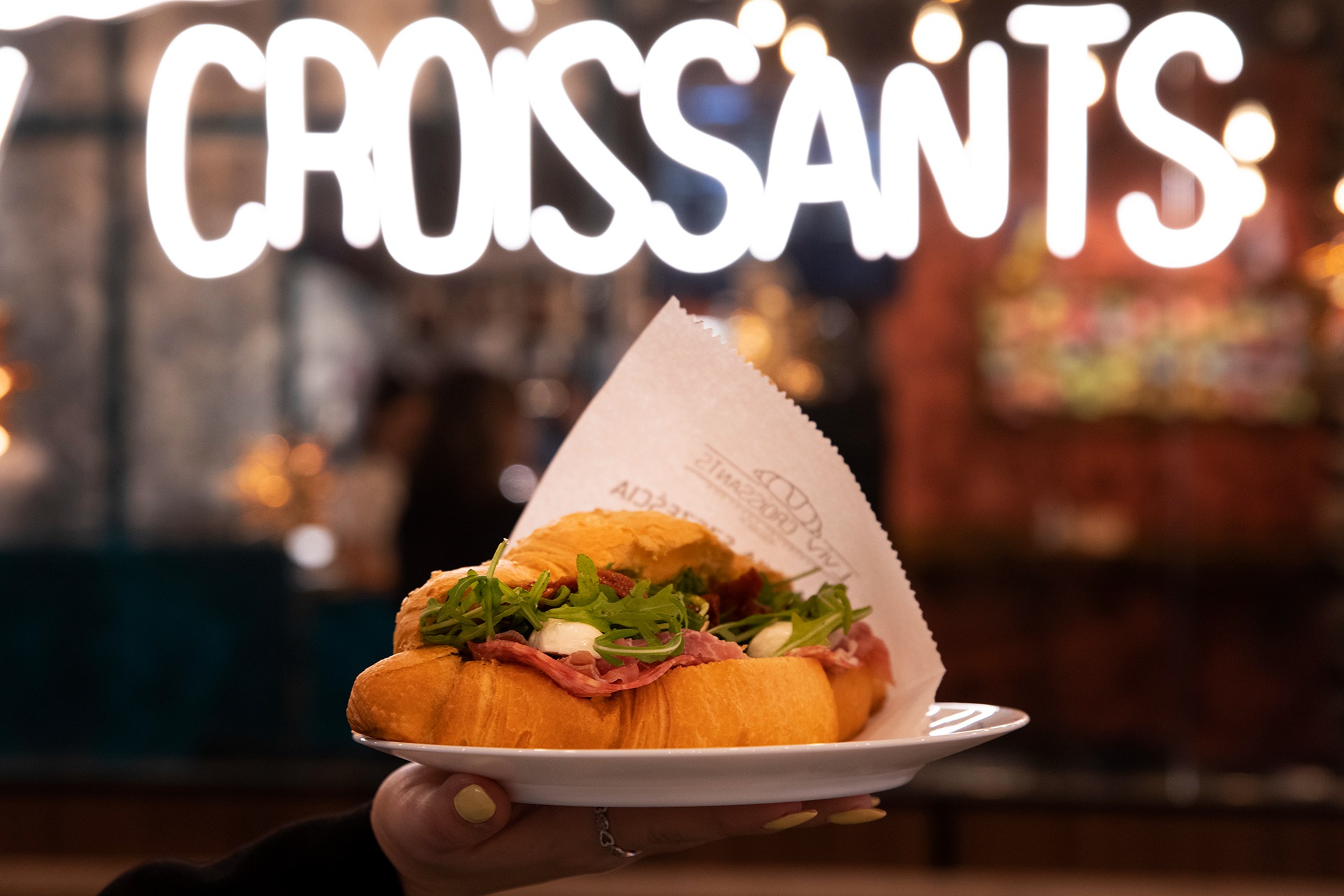 Kadr z bliska: ręka trzyma biały talerz z croissantem wypełnionym szynką, rukolą i mozzarellą na tle neonowego napisu 'CROISSANTS' i rozmytego wnętrza kawiarni.