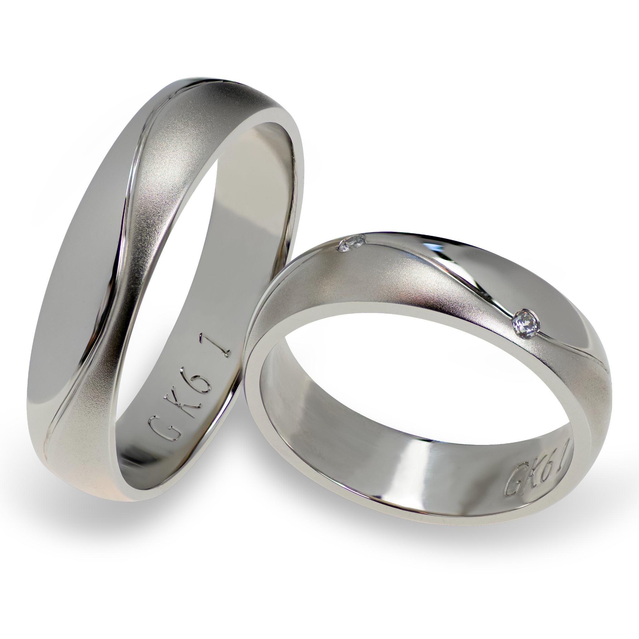 Dwa srebrne, matowe obrączki ślubne z wygrawerowanym napisem GK61 i pojedynczym diamentem na jednej z nich, leżące na białym tle.