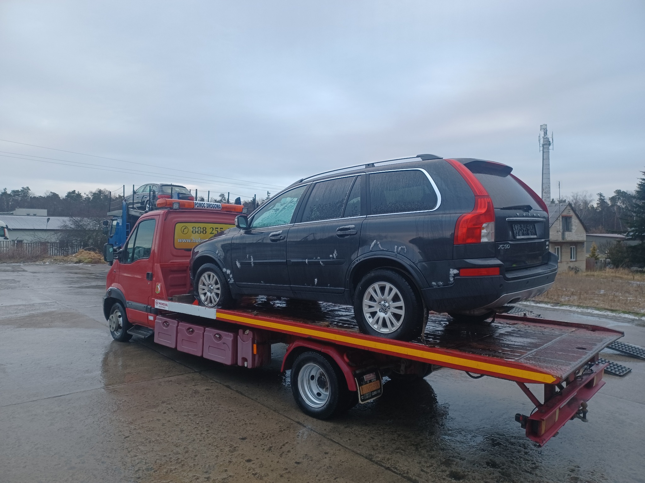 Czerwona laweta z załadowanym czarnym Volvo XC90 na mokrym placu, widoczna wieża telekomunikacyjna w tle.