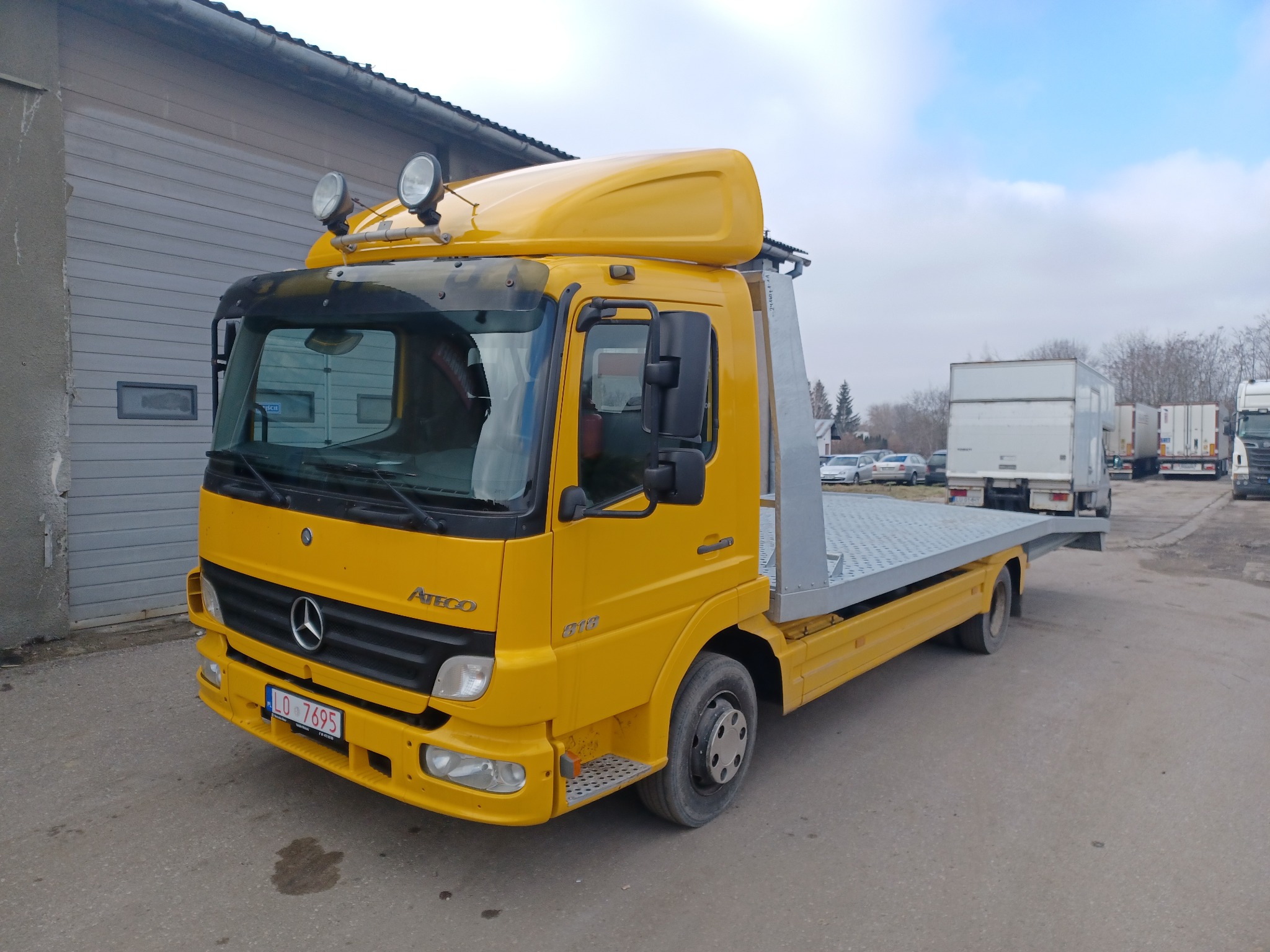 Żółty samochód ciężarowy Mercedes-Benz Atego z platformą lawetową, zaparkowany na betonowym placu, z dodatkowymi lampami na dachu kabiny.