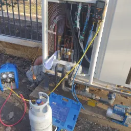 Otwarta obudowa pompy ciepła z widocznymi rurami miedzianymi i przewodami elektrycznymi, podłączona do manometru elektronicznego; obok butla z gazem, niebieska pompa próżniowa i narzędzia na ziemi.