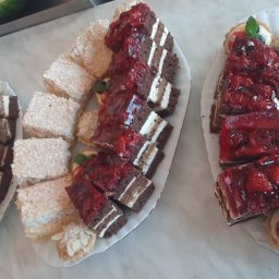 CATERING TRADYCJA PATRYCJA JASIŃSKA - Dwa podłużne talerze z różnorodnymi ciastami: czekoladowe z wiśniami, kokosowe kwadraty, kruche babeczki z kremem i owocami, oraz kawałek arbuza w tle.