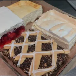 Kartonowe pudełko z czterema różnymi kawałkami ciast: ciasto z galaretką i bitą śmietaną, sernik, ciasto z bezą i ciasto makowe z lukrem w kratkę.